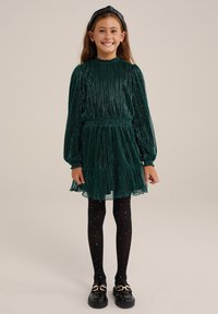 Tummanvihreä laskostettu mekko pitkillä hihoilla ja smokkivyötäröllä, kerroksellinen hame, paritettuna mustien tähtikuvioisten sukkahousujen ja mustien kenkien kanssa.