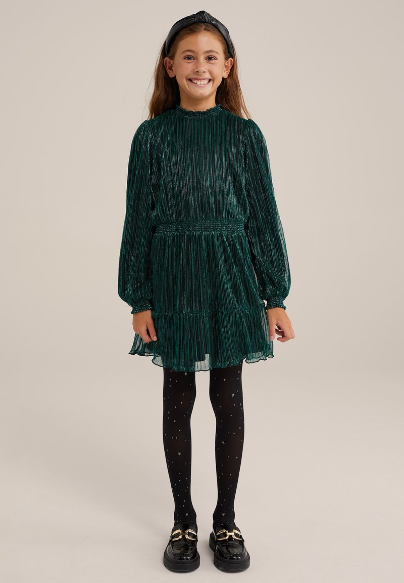 Tummanvihreä laskostettu mekko pitkillä hihoilla ja smokkivyötäröllä, kerroksellinen hame, paritettuna mustien tähtikuvioisten sukkahousujen ja mustien kenkien kanssa.