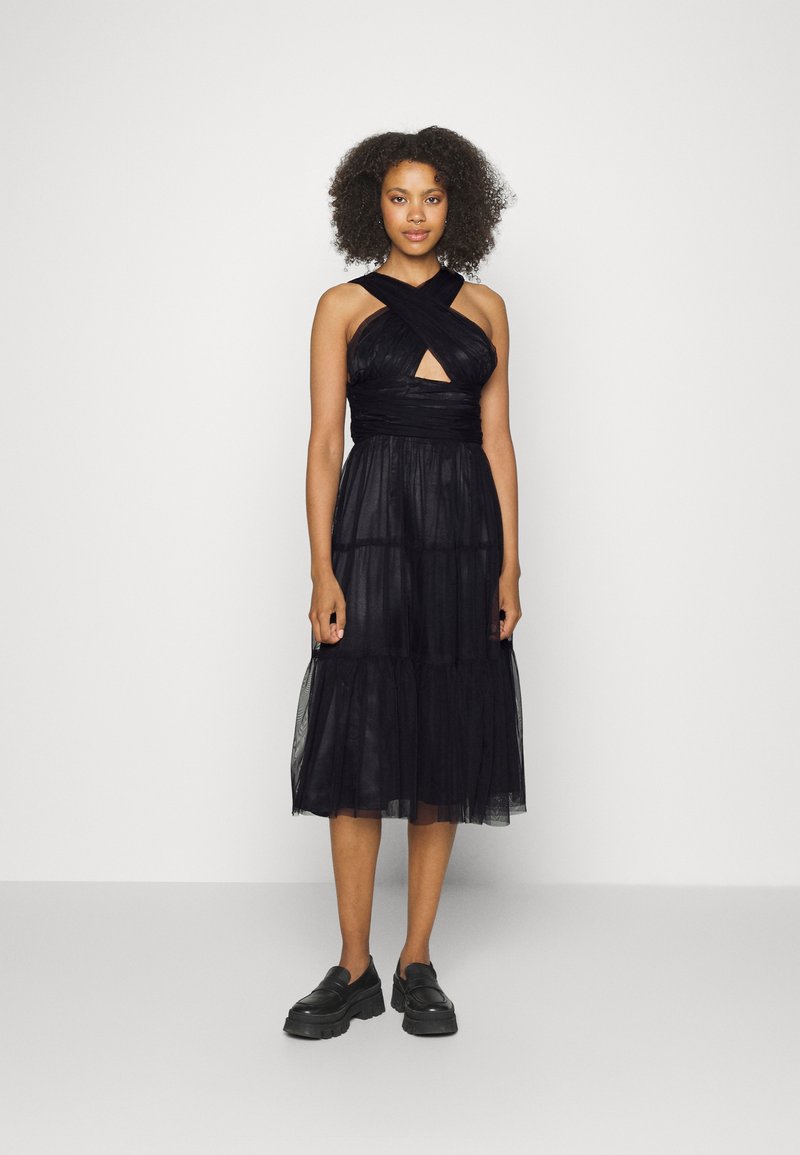 Vestido preto sem mangas com um corpete ajustado, alças cruzadas e uma saia em camadas e translucida. Estilizado com mocassins pretos chunky.