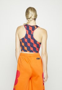 Una donna con capelli biondi intrecciati indossa un top corto senza maniche a righe ondulate rosse e blu navy e pantaloni larghi arancioni brillanti con forme rosse.