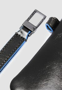 Correa de cuero negro con costuras y detalles en azul, con una hebilla rectangular metálica. Textura suave y diseño limpio.