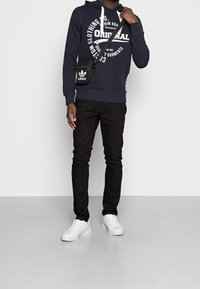 Granatowy hoodie z białym graficznym napisem, czarne dżinsy i białe sneakersy. Mała czarna torba na ramię z białym logo Adidas.