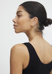 Melna tank topa ar gludu faktūru, apaļš kakla izgriezums, modeļa mati sakrauti bulciņā, valkā nelielas auskaru tapas, skats no sāniem pret baltu fonu.