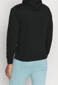 Felpa nera con finitura testurizzata, dotata di cappuccio regolabile con lacci, maniche lunghe e polsini a costine. Abbinata a pantaloni azzurri chiari.