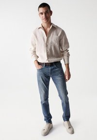 Camisa de lino de botones en beige claro, jeans ajustados azul y zapatillas beige; diseño minimalista con un bolsillo en el pecho y un ligero desgaste.