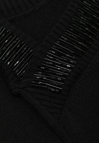 Detailný záber čiernej pletennej látky s lesklými čiernymi cylindrickými korálkami prišitými v paralelných radoch, ktoré tvoria dekoratívny vzor.