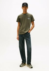 Camiseta de algodón verde oliva con un logo blanco, combinada con jeans de mezclilla azul y zapatillas deportivas grises con acentos rojos y negros.