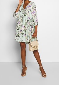 Sukienka w kwiatowy wzór w miętowym kolorze, z rękawami o długości trzech czwarte oraz dekoltem w serek. Uzupełniona o małą, teksturowaną, beżową torebkę oraz sandały na paski.