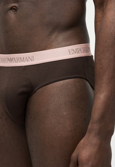 Άνδρας κορμός και άνω μηρός φορώντας σκούρα καφέ σλιπ Emporio Armani με μπεζ ζώνη μέσης σε λευκό φόντο.