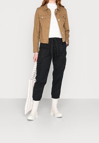 Ruskea denim-takki, valkoinen ribbipaita, mustat verkkopantsit nyörillä ja valkoiset nilkkasaappaat. Raitakassi. Minimalistinen muotoilu.