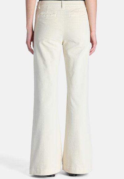 Personne portant un pantalon large en velours côtelé couleur crème, debout de dos sur un fond uni.