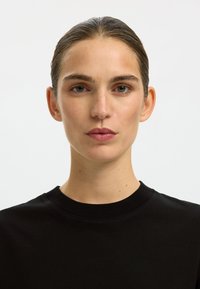 Črna bombažna majica z okrolim izrezom. Ima gladko teksturo in prilegajoč se kroj. Brez vidnih vzorcev ali logotipov.