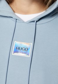 Sudadera de color azul claro que presenta un tejido suave, capucha con cordones y un parche iridiscente que muestra el texto "HUGO" en fuente negra.