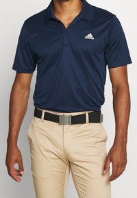 Osoba má na sobě tmavě modrou polo košili s logem Adidas, béžové kalhoty a černý opasek, pózující s neutrálním výrazem.