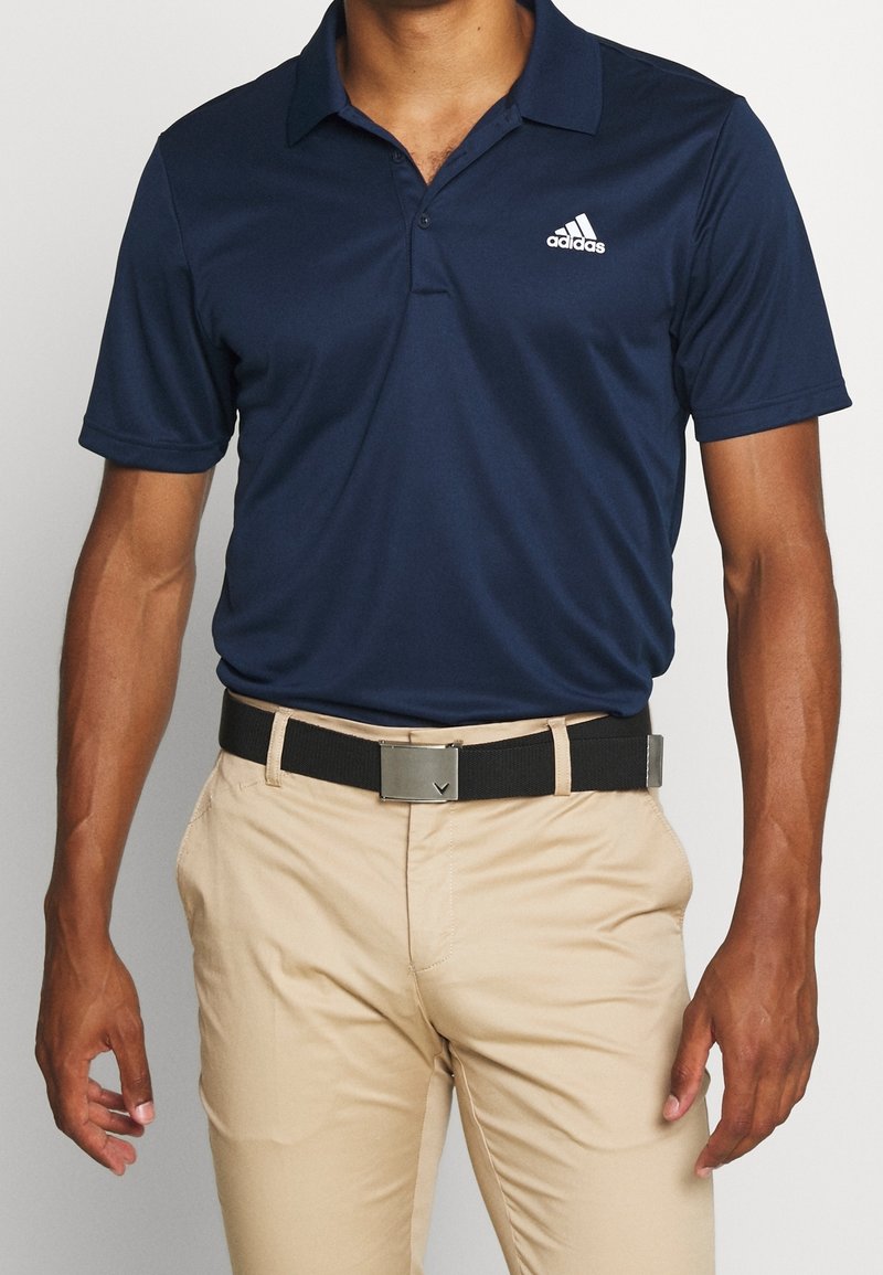 Osoba má na sobě tmavě modrou polo košili s logem Adidas, béžové kalhoty a černý opasek, pózující s neutrálním výrazem.