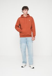 Πορτοκαλί φούτερ με τσέπη καγκουρό και κορδόνια, συνδυασμένο με ανοιχτό μπλε τζιν και λευκά αθλητικά παπούτσι α. Απλός σχεδιασμός, χωρίς ορατά σχέδια.