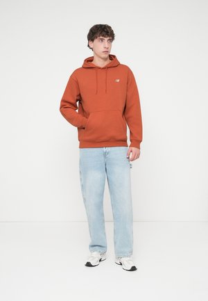 Oransje hettejakke med kengurulomme og snorer, kombinert med lyseblå jeans og hvite joggesko. Enkel design uten synlige mønstre.