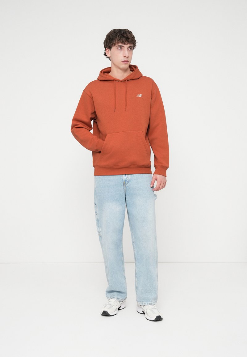 Oranger Kapuzenpullover mit Kängurutasche und Kordelzug, kombiniert mit hellblauen Jeans und weißen Turnschuhen. Einfaches Design, keine sichtbaren Muster.