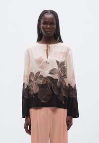 Blouse fluide rose clair avec motifs floraux dans des teintes plus foncées, dotée d'une encolure en trou de serrure et de manches longues. Associée à un pantalon rose clair.