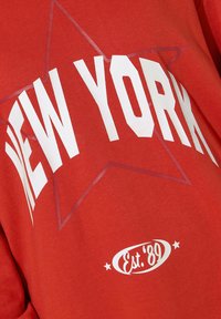 Felpa rosso con grande scritta bianca che recita "NEW YORK" e un design a stelle. Include un logo circolare con "Est.'89" e piccole stelle.