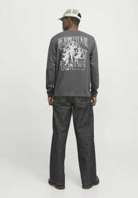 Langärmliges graues Shirt mit grafischem Text auf der Rückseite "WORLD TOUR SOLD OUT" und detaillierten Illustrationen; kombiniert mit weit geschnittenen dunklen Jeans.