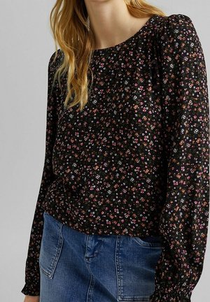 Femme portant un chemisier noir à manches longues avec de petites fleurs multicolores et un jean en denim bleu, vue du cou jusqu'aux hanches.