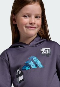 Smiling young girl with red hair wearing a purple hoodie featuring pixelated pandas and a blue pixelated triangular design.