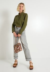 Top de manga larga verde oliva con cuello alto, combinado con vaqueros de pierna ancha gris claro y sandalias de plataforma marrones. Bolso de mano con un patrón multicolor.