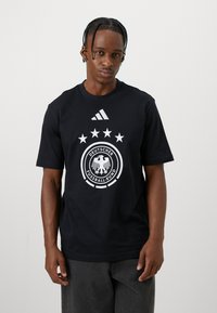 T-shirt de algodão preto com um logo branco da Associação Alemã de Futebol, cinco estrelas acima do logo e um logo da Adidas no canto superior esquerdo.