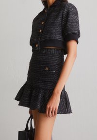 Svart tweed-set med två delar som består av en kort jacka med guldfärgade knappar och korta ärmar, samt en utsvängd kjol med matchande detaljer.