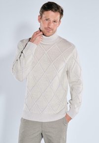 Cremfarbener Rollkragenpullover aus Strickstoff, mit einem Rautenmuster und gerippten Bündchen und Saum, getragen über grauen Hosen.