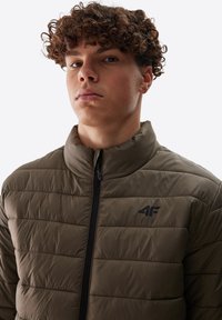 Chaqueta acolchada verde oliva con cuello alto, patrón de costura horizontal, cierre frontal con cremallera y un pequeño logo negro en el pecho.
