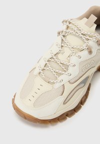 Zapatilla deportiva en crema y beige con paneles de malla, superposiciones texturizadas, cordones con detalles reflectantes y una suela de goma con un patrón de panal.