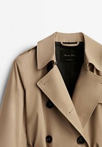 Trench-coat beige avec de gros boutons noirs. Il présente un large col, des épaulettes et une texture de tissu lisse et léger. Étiquette intérieure visible.