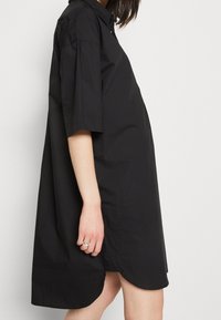 Abito nero a camicia, maniche corte, vestibilità casual. Presenta un colletto e spacco laterale. Tessuto morbido e leggero.