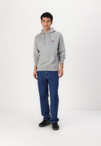 Hanorac din bumbac gri cu buzunar frontal, snur ajustabil și patch cu logo. Asortat cu blugi din denim albastru și încălțăminte neagră.