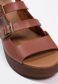 Sandale compensée en cuir marron avec deux brides réglables à boucle métallique et semelle intérieure rembourrée couleur beige sur fond blanc.