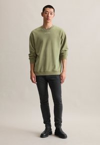 Jonge man in een olijfgroen sweatshirt, zwarte slim fit jeans, zwarte laarzen, zilveren ketting en armband, staand tegen een effen beige achtergrond.