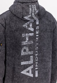 Black hoodie made of soft fabric, featuring a distressed texture. "ALPHA INDUSTRIES" printed vertically in faded white on the back.