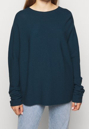 Personne portant un pull ample bleu foncé à manches longues avec un jean bleu clair sur un fond clair uni.