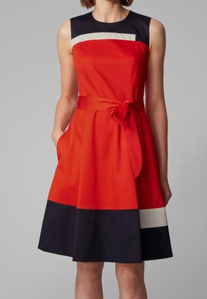 Bez rękawów sukienka do kolan z czerwonym górnym stanikiem i spódnicą, czarno-białe poziome paski oraz przewiązany pasek w talii, noszona przez stojącą osobę.