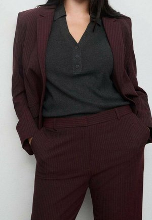 Femme portant une chemise gris foncé boutonnée, rentrée dans un costume à rayures fines bordeaux, les mains dans les poches, devant un mur uni.