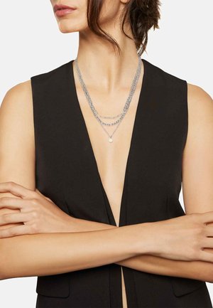 Zilveren multi-laags ketting met schakels en een kleine parelaccent, gedragen over een zwarte top met een diepe v-hals.