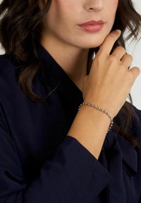 Bracciale di perline in argento su un polso, che mostra perline metalliche arrotondate con una finitura liscia. La mano è posizionata vicino a un volto che indossa abiti scuri.