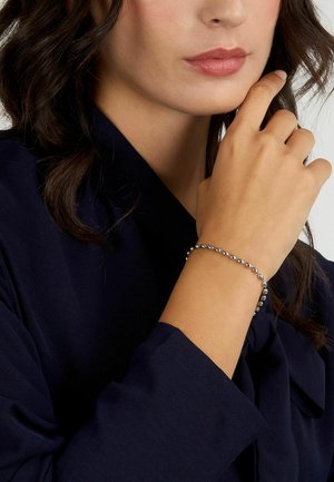 Bracciale di perline in argento su un polso, che mostra perline metalliche arrotondate con una finitura liscia. La mano è posizionata vicino a un volto che indossa abiti scuri.