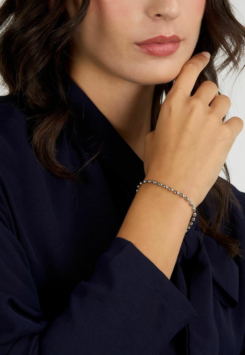 Bracciale di perline in argento su un polso, che mostra perline metalliche arrotondate con una finitura liscia. La mano è posizionata vicino a un volto che indossa abiti scuri.