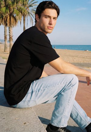 Mladý muž v černém tričku a světlých džínách sedí na kamenné lavičce u pláže s palmami pod jasně modrou oblohou.