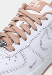 Baskets Nike Air Force 1 en cuir blanc avec des lacets beige, un label beige sur la languette, des accents beiges cousus et un petit swoosh Nike en argent près de la semelle.