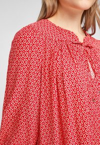 Blouse rouge avec un motif floral blanc, présentant un décolleté froncé et un détail à nouer. Tissu léger et coupe ample.