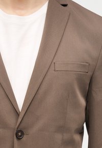 Καφέ ραμμένο σακάκι με V γιακά, τσέπη στο στήθος και ένα μοναδικό σκούρο κουμπί, φορεμένο πάνω από λευκό μπλουζάκι με στρογγυλή λαιμόκοψη.